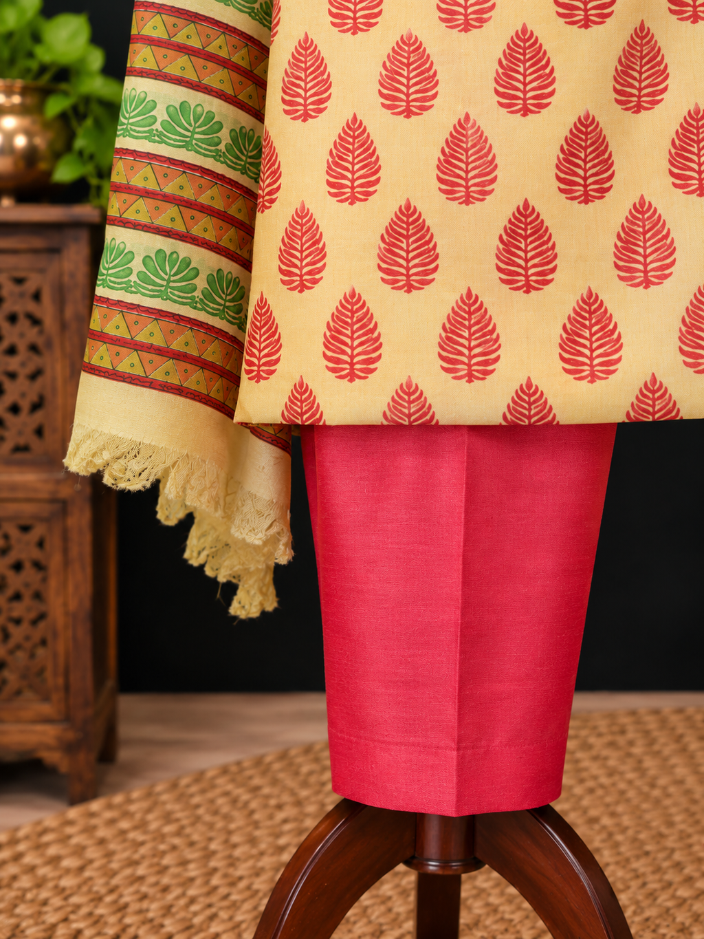 A close-up of a traditional outfit draped on a wooden stand, featuring a light beige kurta with red leaf block prints, a bright pink skirt, and a patterned cream dupatta with green and red motifs.