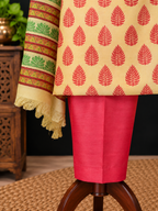 A close-up of a traditional outfit draped on a wooden stand, featuring a light beige kurta with red leaf block prints, a bright pink skirt, and a patterned cream dupatta with green and red motifs.