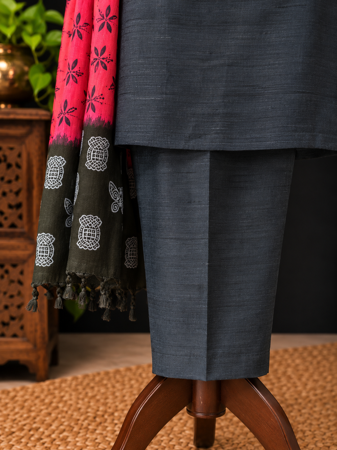 A close-up of a dark slate gray three-piece silk suit displayed on a mannequin stand, featuring a matching solid lapel pin and a vibrant pink and black patterned dupatta draped over the shoulder.