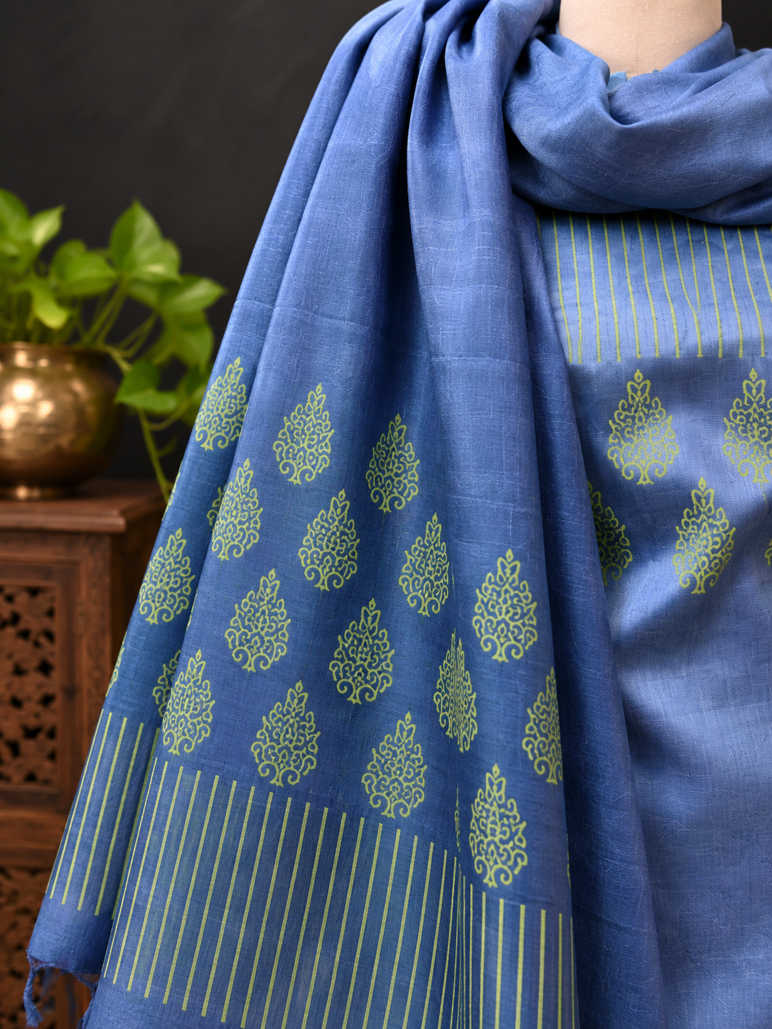 A blue dupatta with yellow-green block-printed floral motifs and striped borders, draped over a model wearing a matching blue printed kurta. A wooden side table with a green plant is visible in the background.