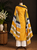 A mustard yellow salwar kameez suit with a patterned dupatta draped on a mannequin, styled in a warm indoor setting with a side table, flowers, and a dark background.