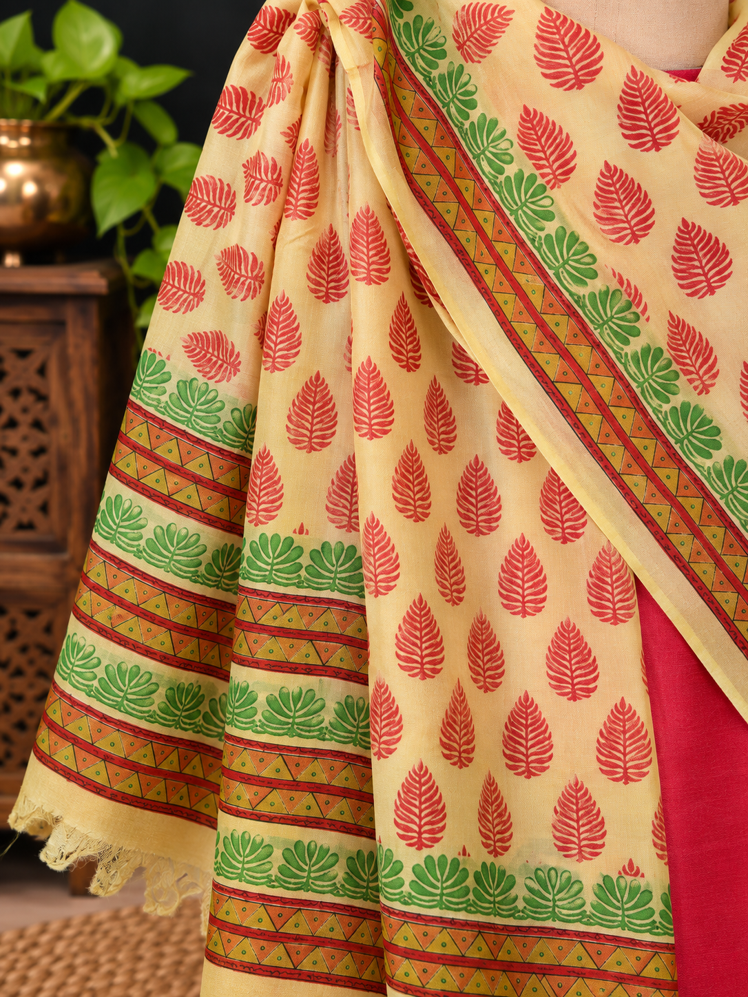 A close-up of a printed fabric silk dupatta lwith red leaf motifs and green-and-red geometric borders, shown against a dark indoor background with a plant and wooden furniture.