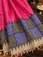 A close-up of a pink fabric textile with a wide decorative border. The border includes blue and beige patterned sections with black ornamental motifs and tassels along the edge.