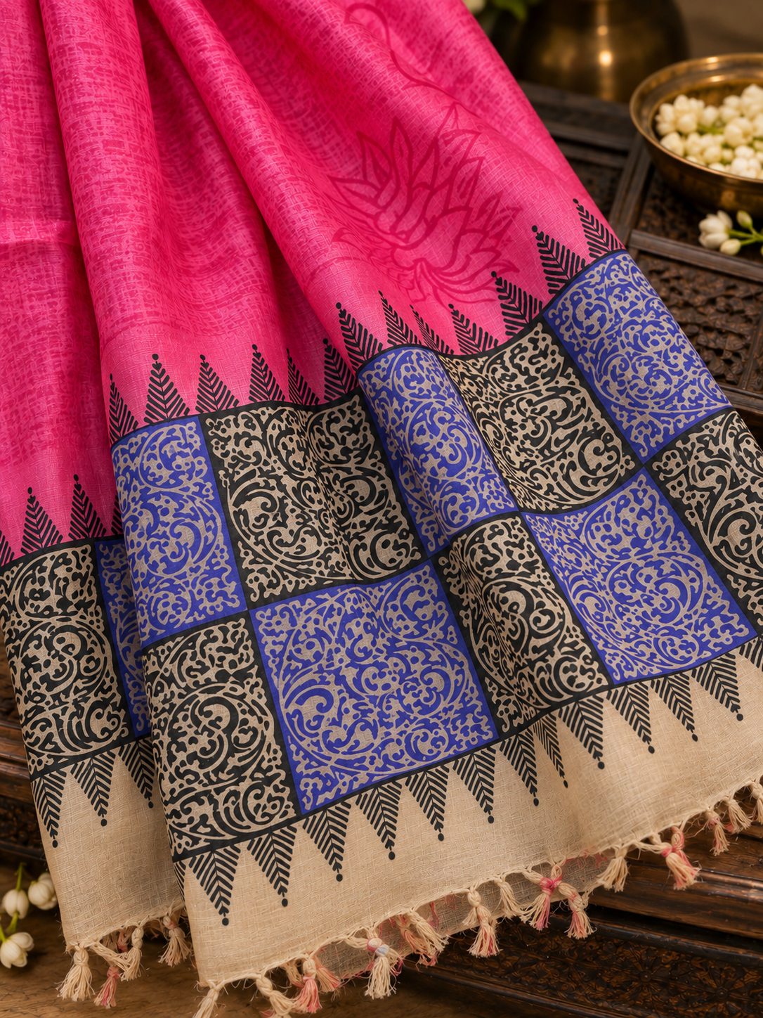 A close-up of a pink fabric textile with a wide decorative border. The border includes blue and beige patterned sections with black ornamental motifs and tassels along the edge.