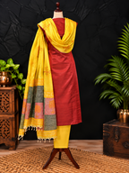 A three-piece women's traditional silk set on a mannequin, featuring a red kurta, yellow trousers, and a yellow dupatta with multicolor patchwork borders and tassels, set against a dark backdrop with decorative plants and wooden furniture.