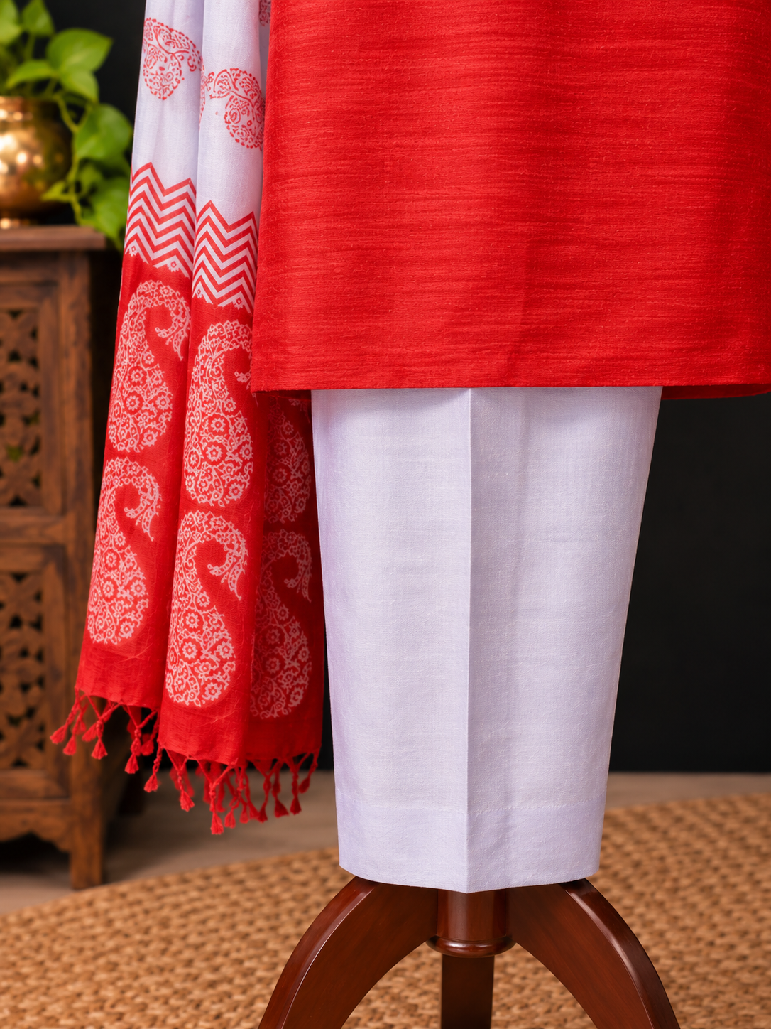 A close-up of a red and white silk traditional outfit displayed on a mannequin stand, featuring a solid red kurta, white trousers, and a red-and-white patterned dupatta with tassels.