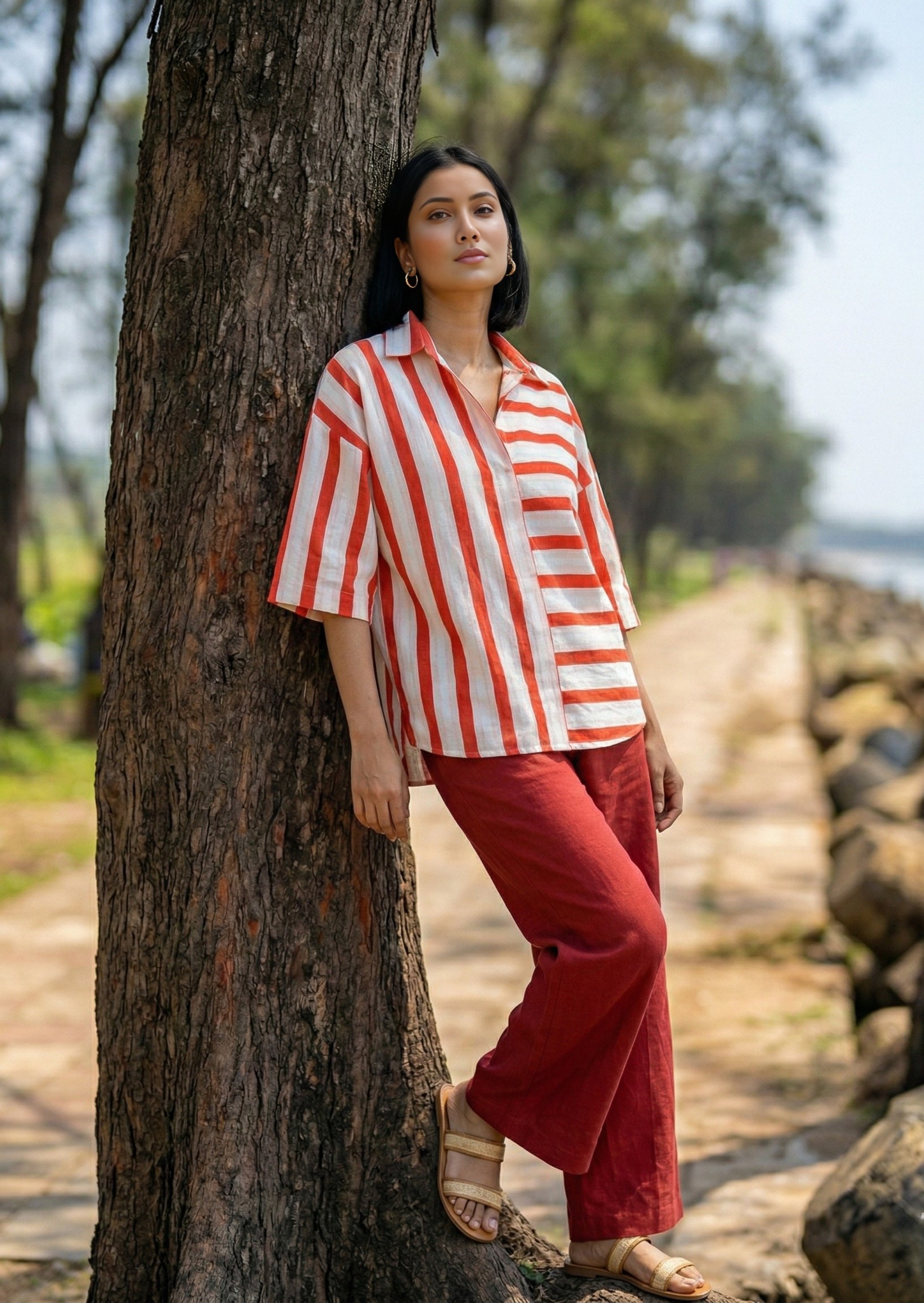 Woman in a red and white striped shirt and red pants leaning against a tree outdoors.