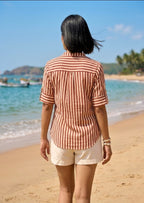 Person wearing a striped shirt and shorts standing on a beach with boats and greenery in the background.