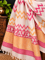 A draped white saree or fabric with pink and orange zigzag patterns, floral borders, and tassels, shown in a styled indoor setting with a wooden cabinet and green plant in the background.