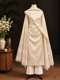 A cream-colored traditional South Asian outfit displayed on a mannequin, featuring a kurta with intricate embroidery, matching trousers, and a draped shawl with geometric patterns and tassels. The mannequin is set against a dark background with decorative plants and a side table.