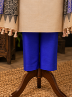 A close-up of a clothing display showing bright royal blue cropped pants paired with a beige tunic and patterned draped fabric with tassels, displayed on a wooden stand over a woven rug.