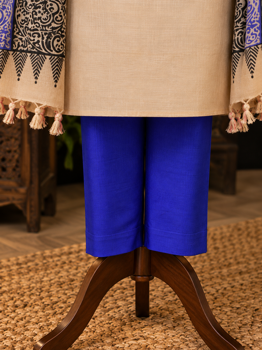 A close-up of a clothing display showing bright royal blue cropped pants paired with a beige tunic and patterned draped fabric with tassels, displayed on a wooden stand over a woven rug.