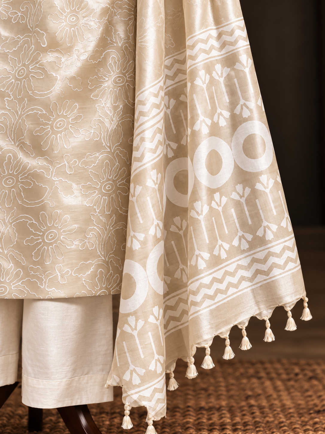 A close-up of a beige and white traditional fabric set, featuring a patterned dupatta with tassels draped over a matching beige garment.